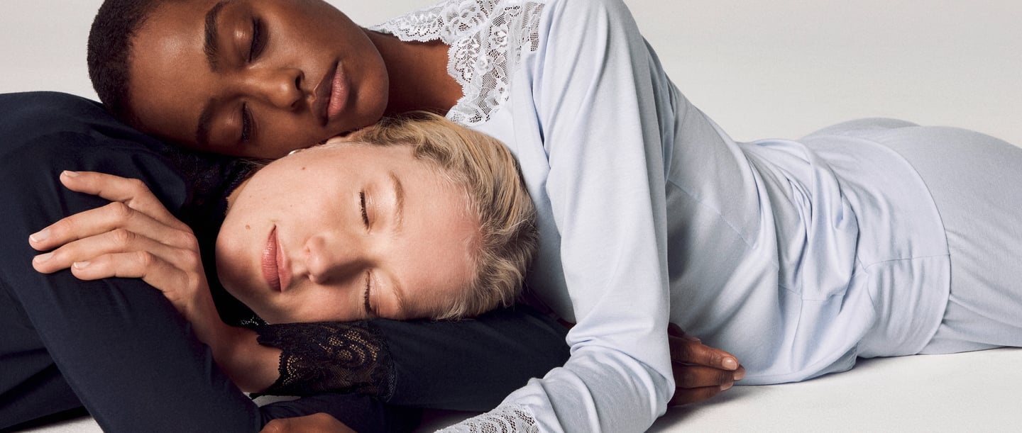 The picture shows two ladies lying close together, their heads resting gently on each other. A white background surrounds them, emphasising their relaxed atmosphere. Both ladies have their eyes closed, which emphasises their calm and serenity.  The ladies are wearing nightwear from CALIDA, whose soft fabric gently envelops their contours. The nightwear exudes elegance and comfort, while its simple beauty complements the image perfectly.