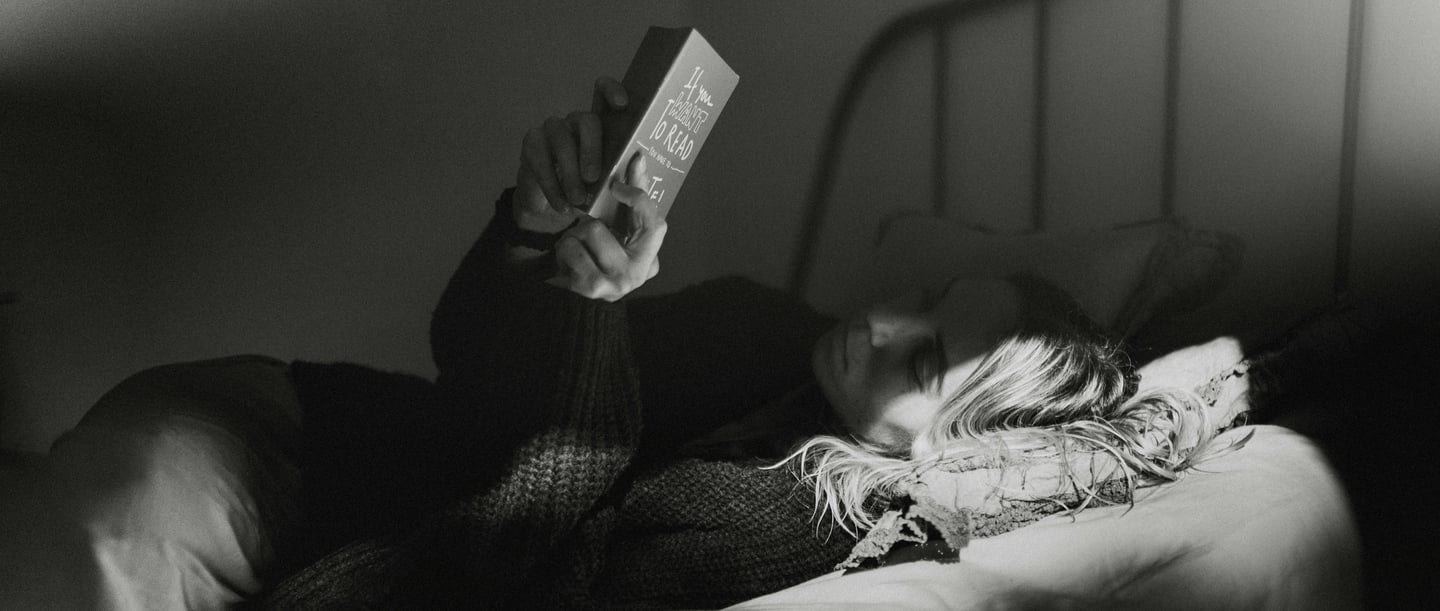 Person lies relaxed in a bed and reads a book in the dim light to calm down and fall asleep better.