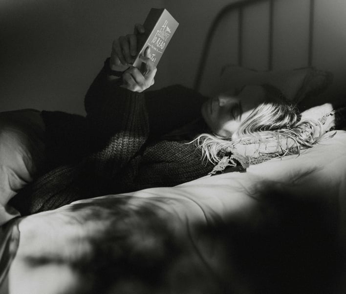Person lies relaxed in a bed and reads a book in the dim light to calm down and fall asleep better.