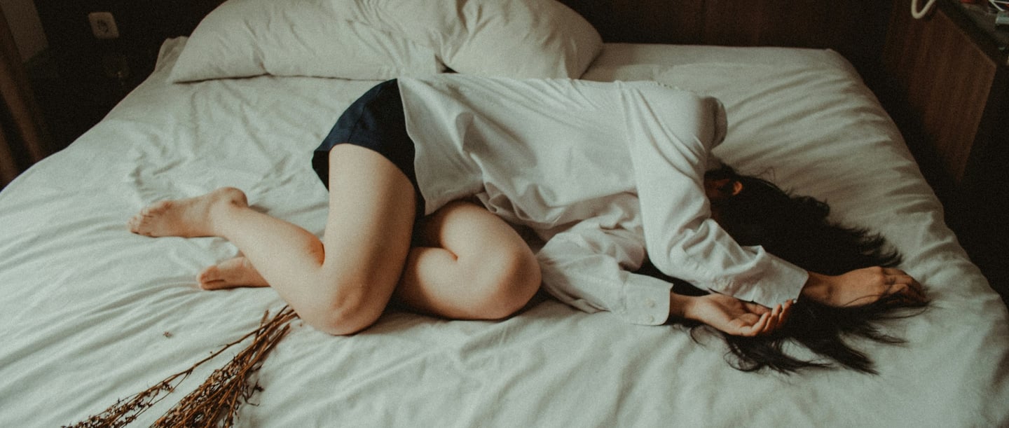 On y voit une femme allongée sur un lit sans oreiller dans une pièce sombre.