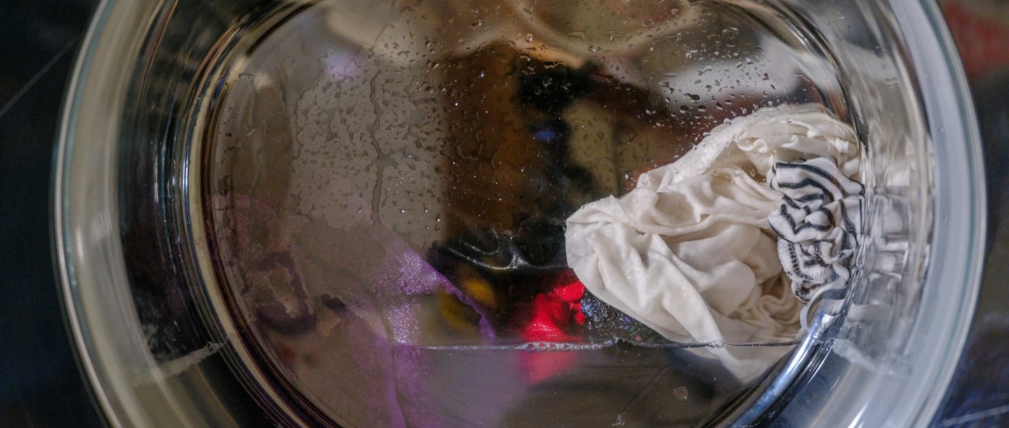 Gros plan sur un tambour de machine à laver dans lequel se trouvent des vêtements mouillés, blancs et colorés. Des gouttes d'eau sont visibles à l'intérieur du tambour.