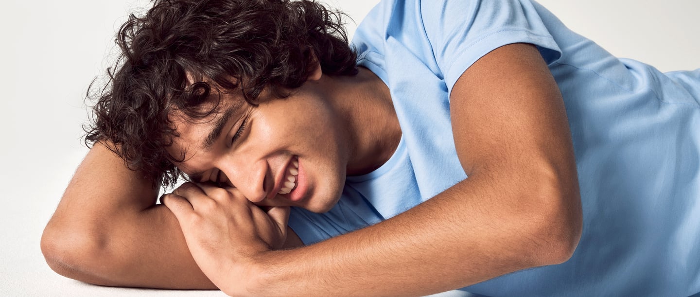 The picture shows a smiling man with curly hair lying on his side on a white background and wearing a light blue T-shirt from CALIDA. He has his eyes closed, symbolising laughter in his sleep.