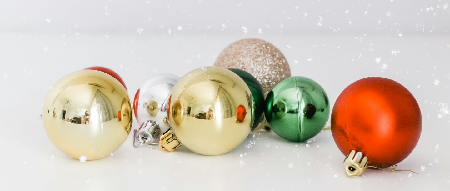 Baubles natalizi in vari colori, tra cui oro, rosso, verde, argento e glitter su sfondo bianco, con un sottile effetto fiocco di neve.