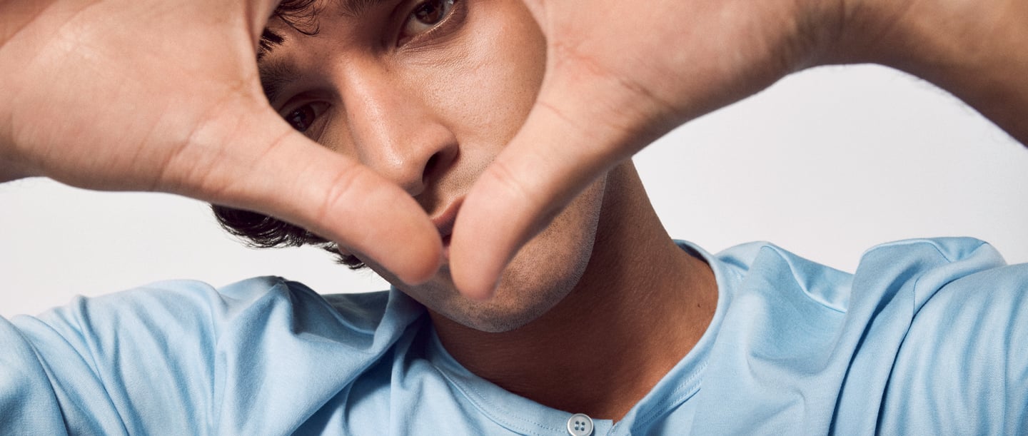 Gros plan sur un jeune homme vêtu d'une chemise bleue, qui tient ses mains devant son visage en forme de cœur. Symbolise la concentration et l'attention, en accord avec le thème des exercices de pleine conscience.