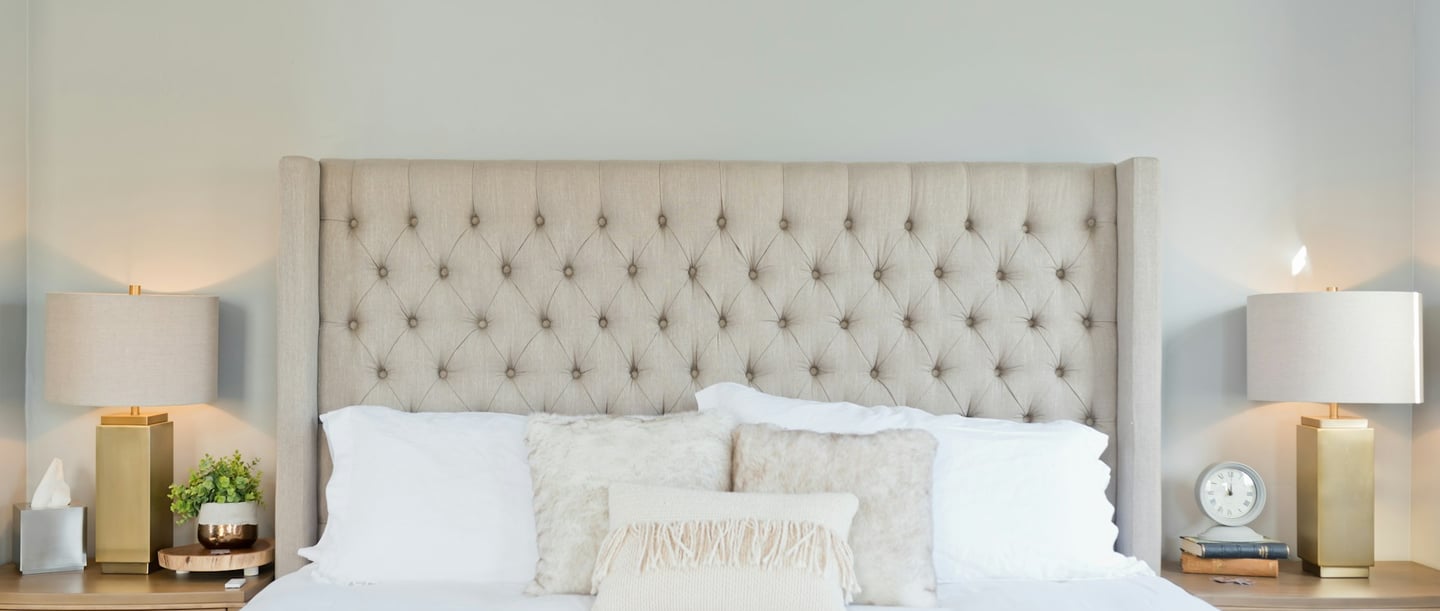 Chambre à coucher lumineuse avec une tête de lit rembourrée beige, deux tables de nuit, des lampes dorées et des draps blancs - symbole de l'air sec dans la chambre à coucher.