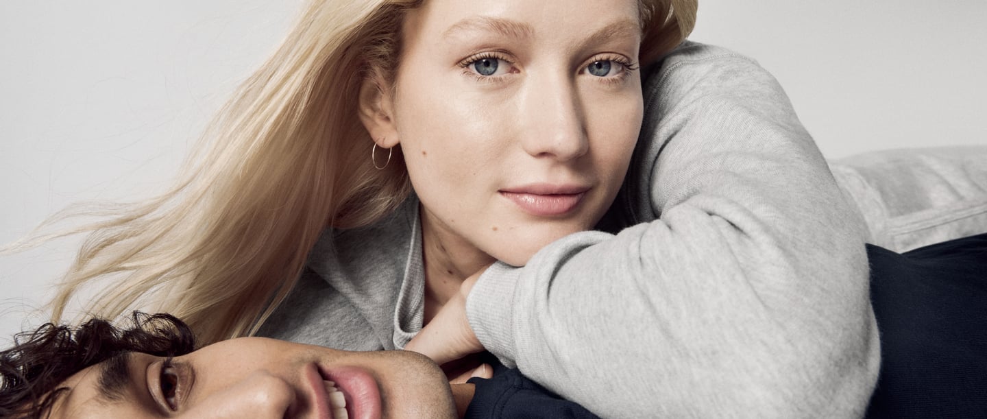 A relaxed couple in CALIDA clothing presenting the onion look. The woman is wearing a grey jumper, the man a dark top, both looking relaxed and stylish.