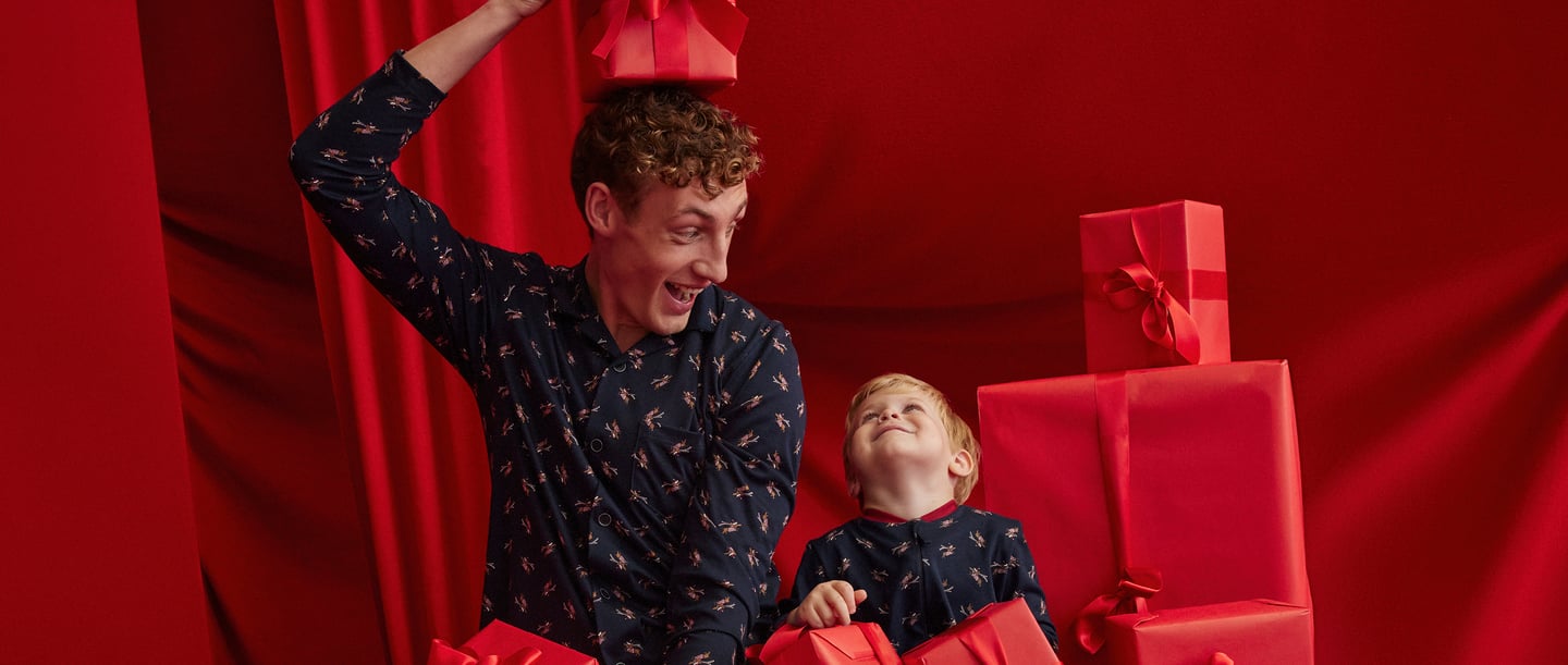Mann und kleiner Junge im Winterpyjama von CALIDA. Der Hintergrund ist ganz rot mit kleinen Geschenken Boxen.