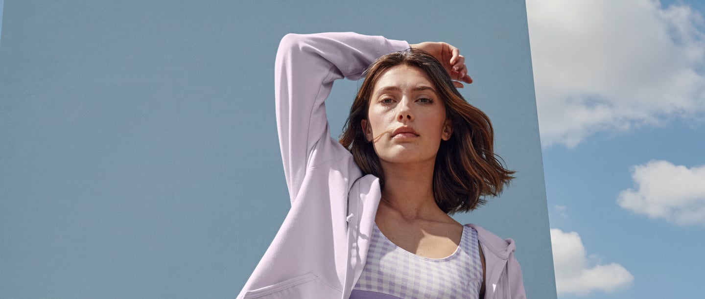 Woman with blue background wears lavender-coloured clothing. Checkered bustier and loungewear jacket by CALIDA from the Elastic Trend and Lounge Tulip series.