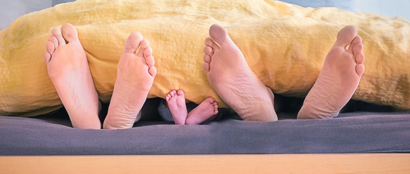 Parents and baby co-sleeping, their feet peeking out from under the duvet together.