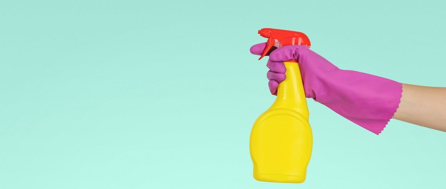 A hand with a pink glove holds a yellow spray bottle with a red lid in front of a turquoise-coloured background.