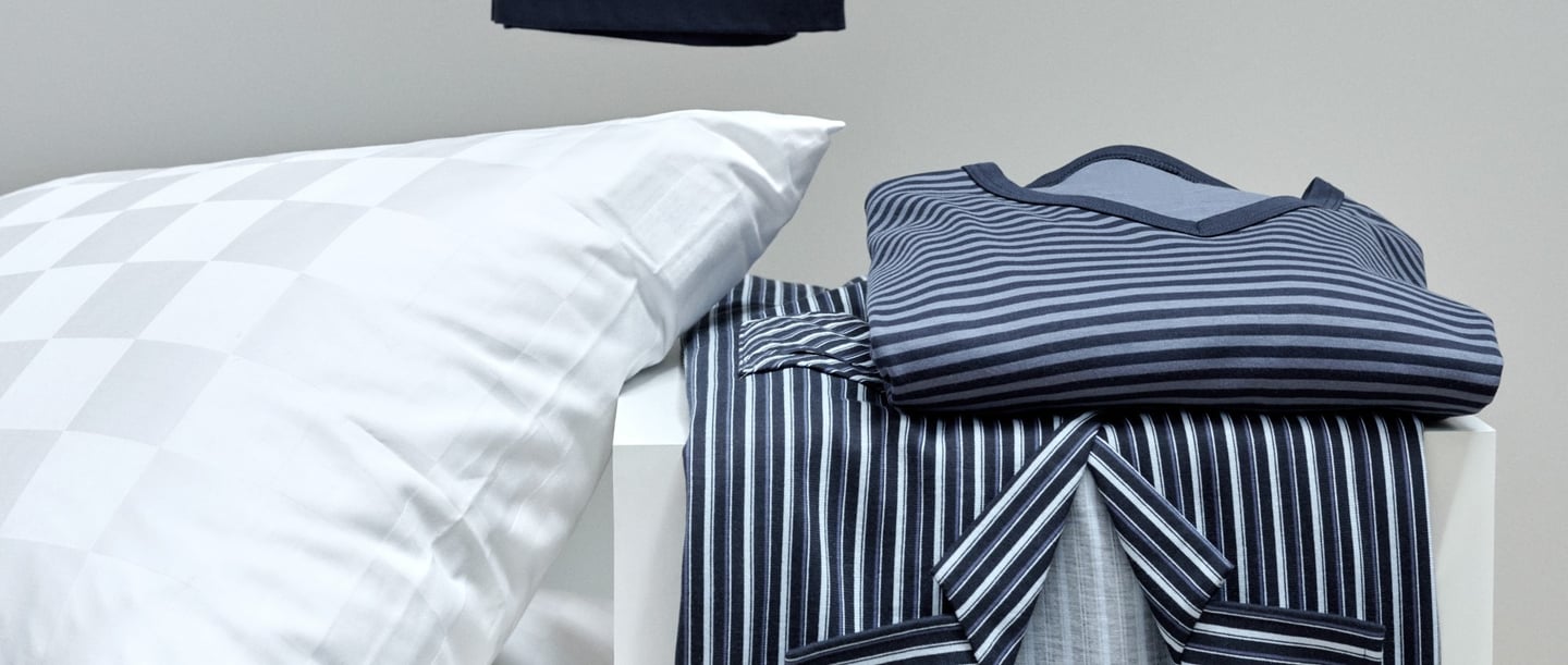 Folded striped pyjamas in shades of blue and grey on a white table next to a white cushion with a subtle checked pattern. A black item of clothing hangs in the background.