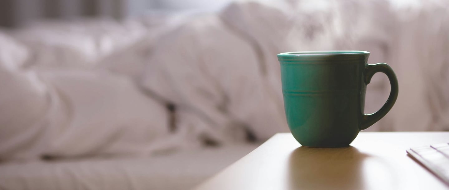 A green cup of steaming coffee stands on a bedside table in front of an unmade bed, symbolising coffee before sleep.