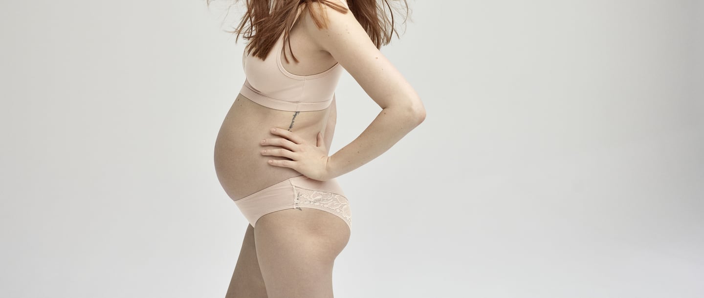 A pregnant woman stands sideways to the camera. Her eyes are closed. She is wearing underwear from CALIDA.
