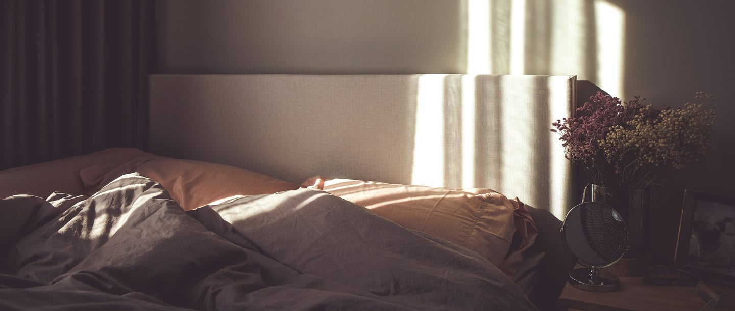 Das Bild zeigt ein Bett mit zwei Kissen und Decken, von einem Sonnenstrahl beleuchtet. Neben dem Bett steht ein Nachttisch mit einem Spiegel, einem Bild und Blumen. Der Raum wirkt gemütlich und einladend.