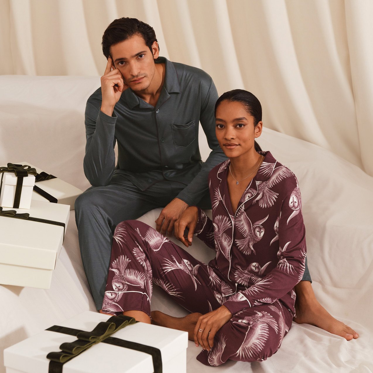 Die Dame trägt den neuen Pyjama der Serie Moonlight Dreams in Weinrot mit Eulen-Print und der Herr den Pyjama der Serie Relax Silk in einem Grauton.