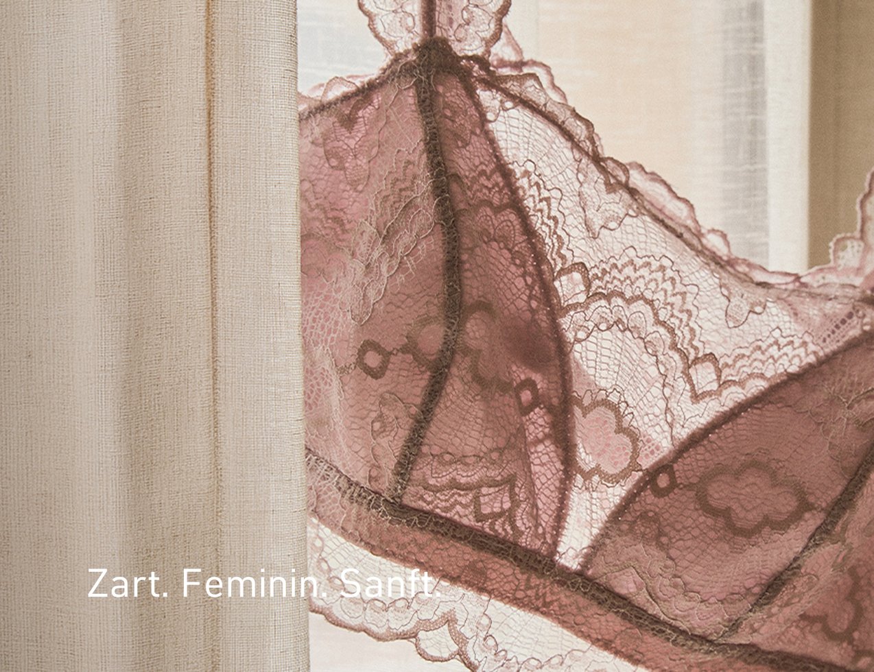 Zarter Spitzen‑BH in Rosé hängt neben einem hellen Vorhang. Die feine Spitze mit floralen Mustern wirkt feminin und sanft 