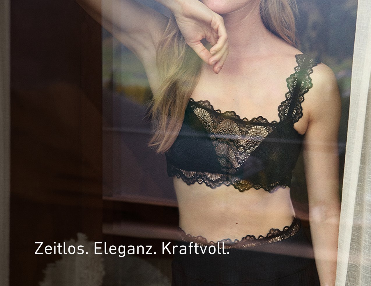 Frau steht hinter einer Glasscheibe und trägt einen schwarzen Spitzen‑BH mit feinen Details