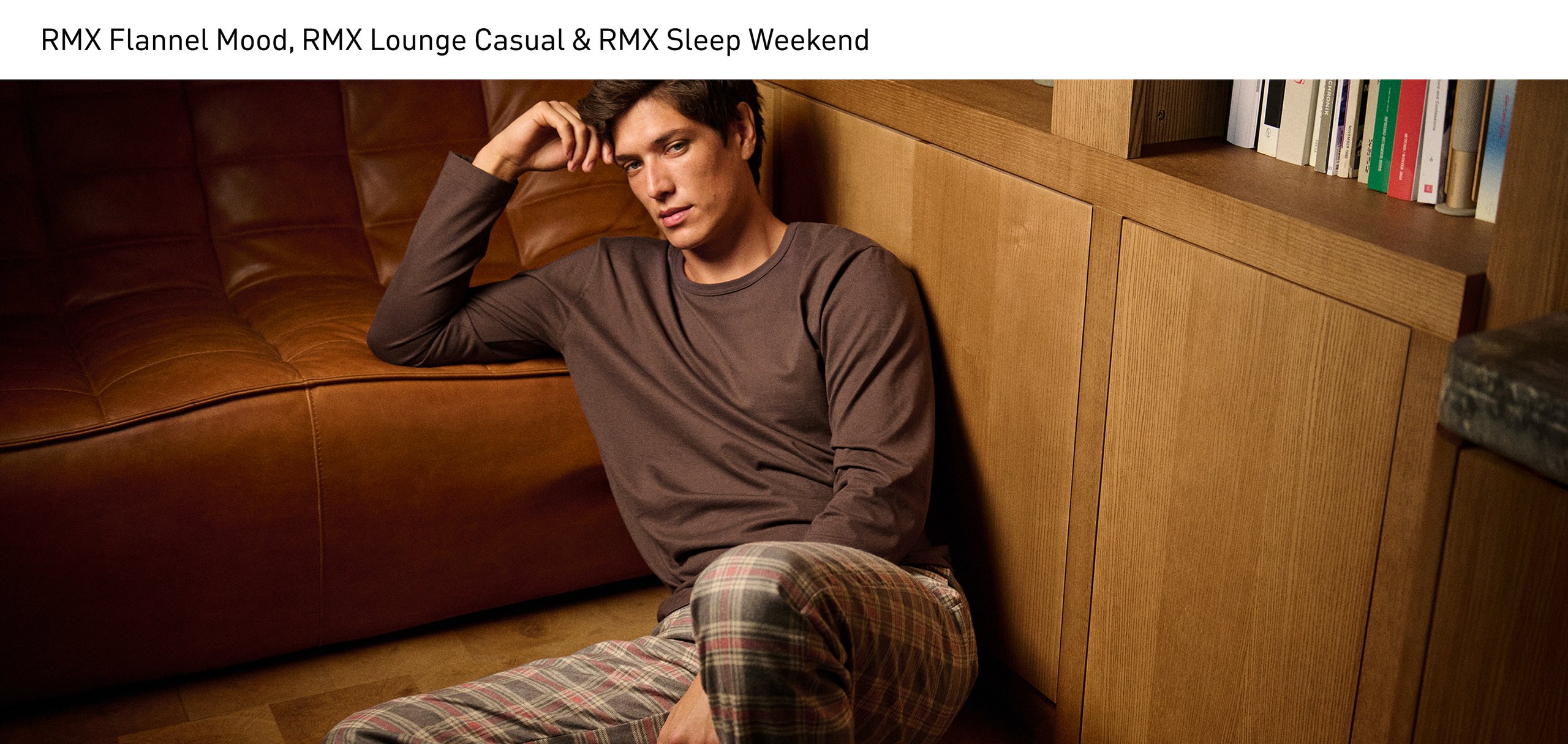 Der Mann sitzt entspannt auf dem Boden und trägt ein braunes Langarm-Shirt der Serie RMX Sleep Weekend, kombiniert mit der karierten Pyjamahose der Serie RMX Flannel Mood mit Seitentaschen.