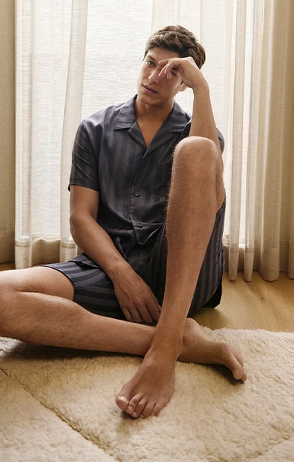 The model is sitting relaxed on the floor, wearing a short button-up pyjama from the Relax Lux series.
