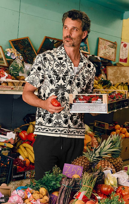 L’uomo si trova in relax al mercato mentre fa acquisti. Indossa una camicia a maniche corte con bottoni della serie RMX Lounge Lecce con stampa moderna per eleganti look mix & match, abbinata a bermuda neri.