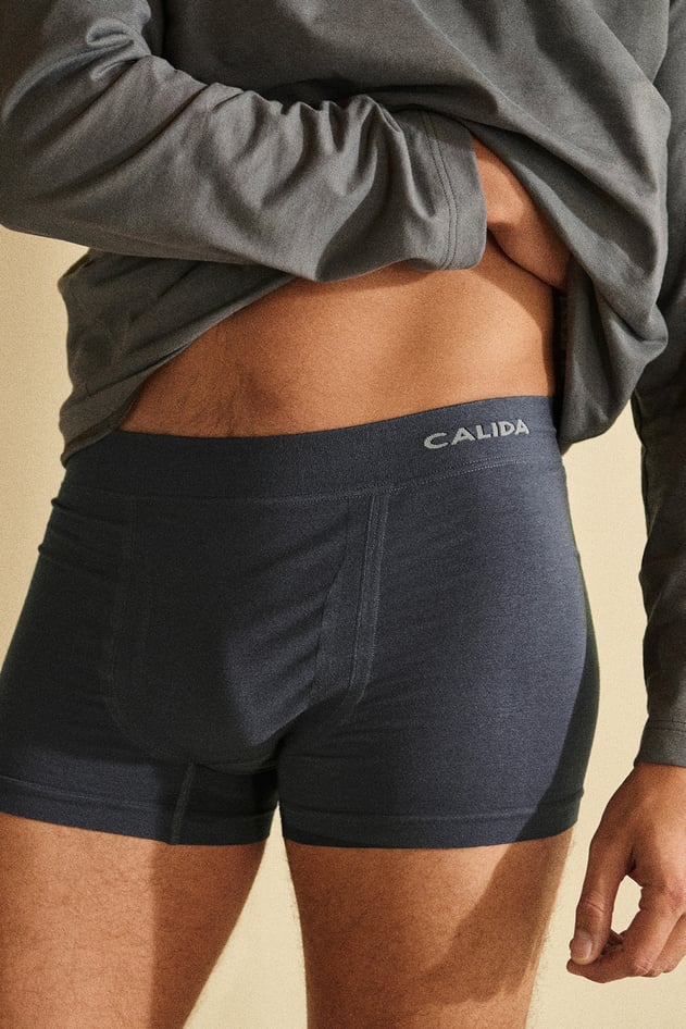 Two pairs of men's boxer shorts by CALIDA in blue and brown folded on a wooden table next to a book.
