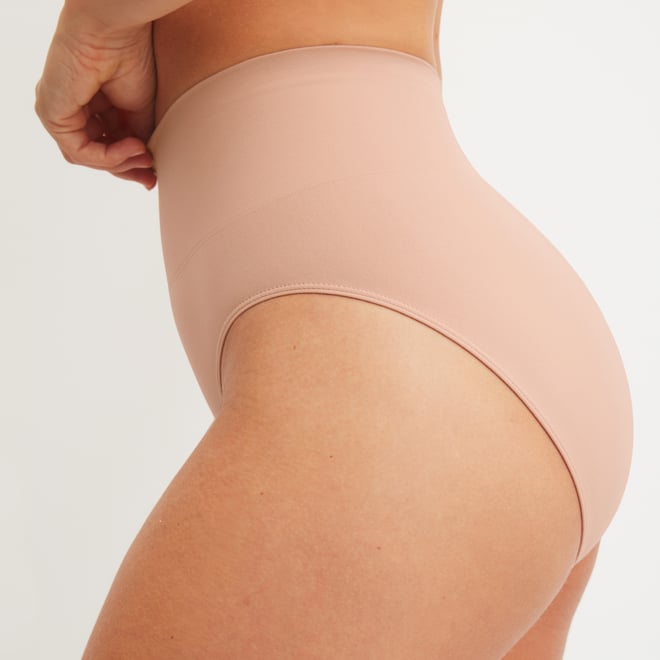 Frau trägt figurformenden Slip in Nude mit hohem Beinausschnitt und glatter Oberfläche für sanfte Formgebung und Komfort – seitliche Rückansicht