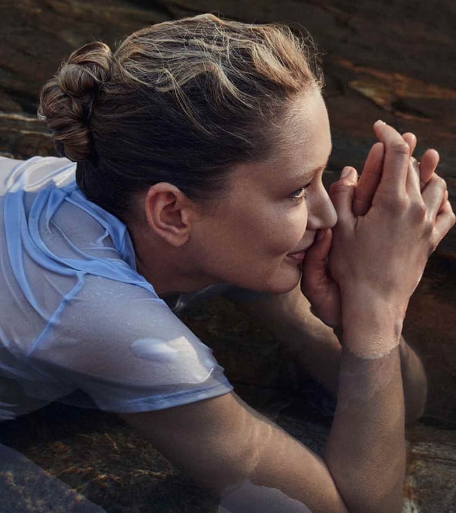 Person lying in shallow water among rocks wearing a wet light blue shirt; calm, nature-connected scene