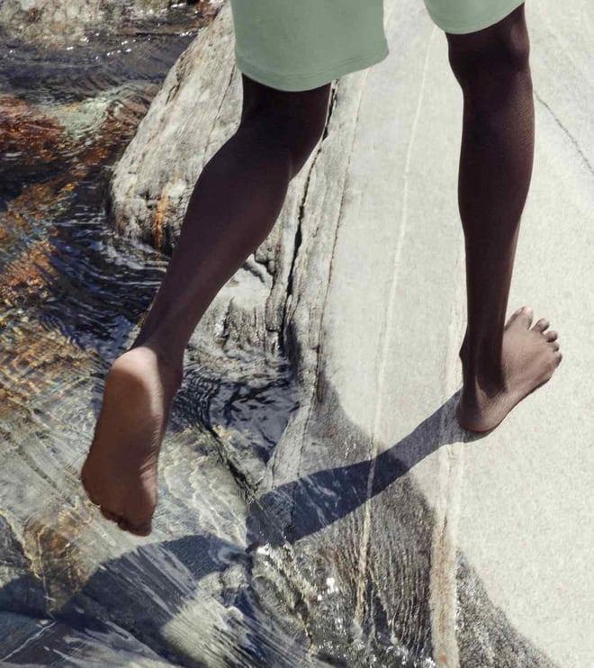 Barefoot person walking on a light rock by the water, focusing on legs and natural surroundings