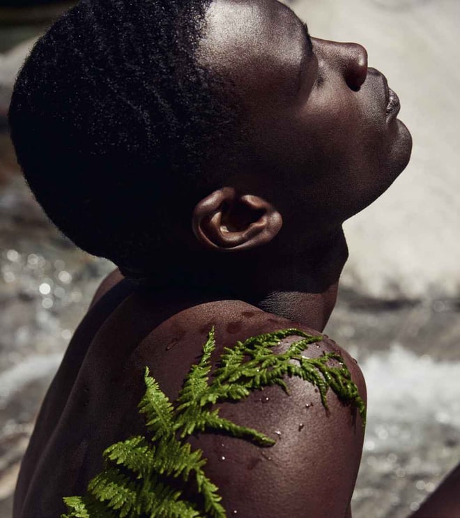 Side close-up of a person with short hair and fern leaves on the shoulder against a natural water landscape