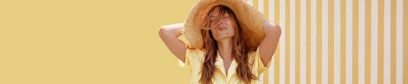 The female model is wearing a yellow striped pyjama top and a straw hat, standing in front of a matching yellow-striped wall.
