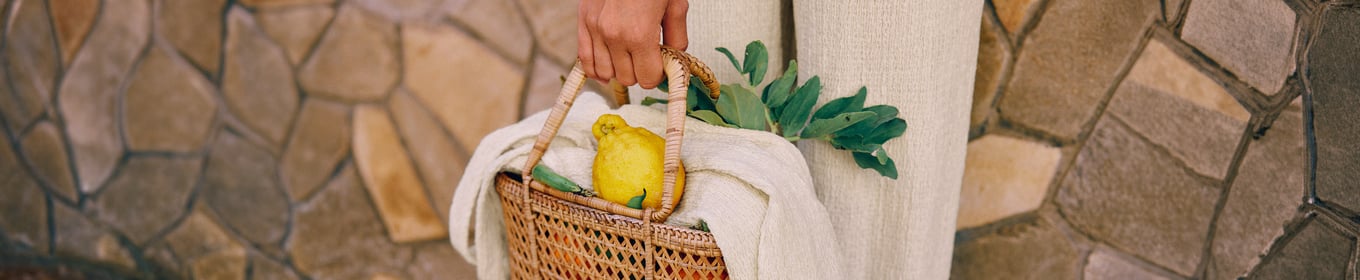 Das Bild zeigt eine Frau in einem schwarzen langen Jumpsiut und einen Mann in einem gemusterten schwarz/weißen Pyjama Oberteil. Beide sind auf einem Markt und haben Tomaten in der Hand.