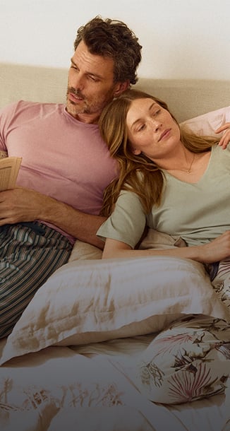 Ein männliches und ein weibliches Model liegen in einem Bett und tragen florale Pyjamas der Marke Calida in Pastelltönen. 