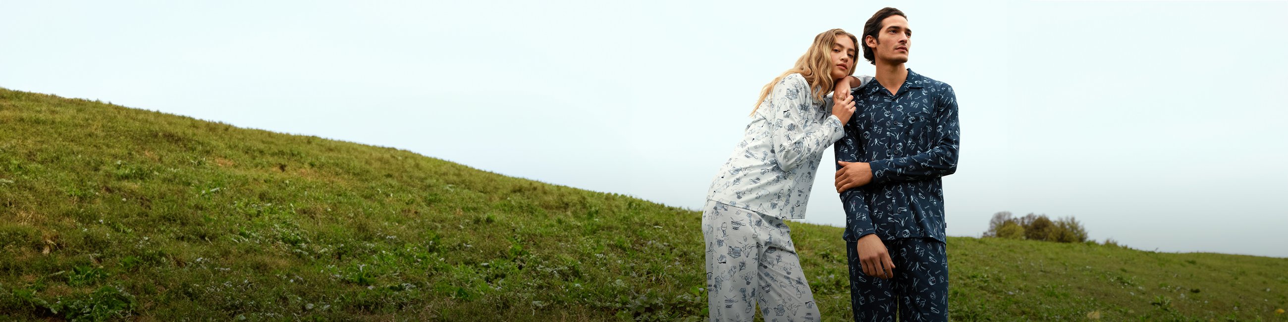 Eine Frau und ein Mann tragen CALIDA Pyjamas mit Print und stehen auf einer Wiese