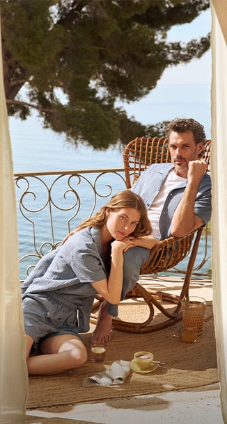 Der Herr sitzt gemütlich in einem Stuhl und schaut verträumt, sie sitzt auf den Boden. Er trägt ein Pyjama aus der Serie RMX Sleep Journey und sie trägt einen Kurzpyjama aus der Serie Favourites Tiles, zusammen ergibt es einen perfekten Partnerlook.