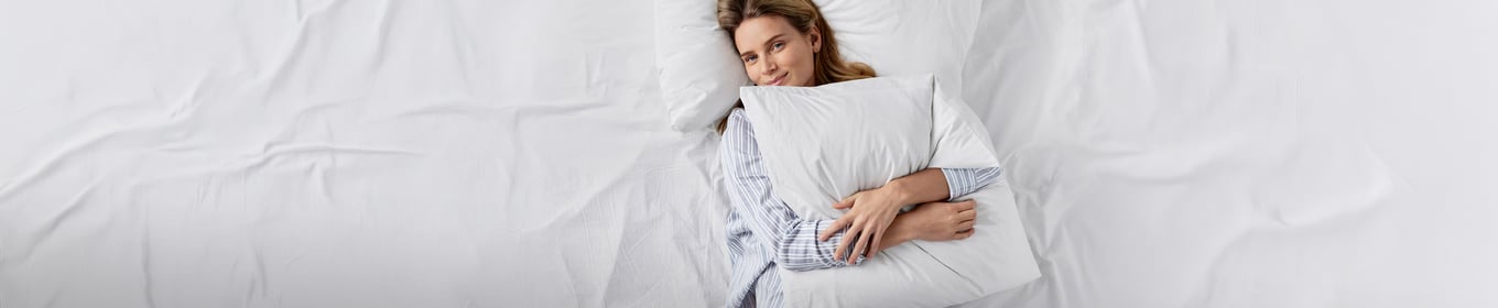 A woman is lying on a white blanket and pillows. She is holding a white pillow in her hands and wearing blue-and-white striped pajamas from CALIDA.
