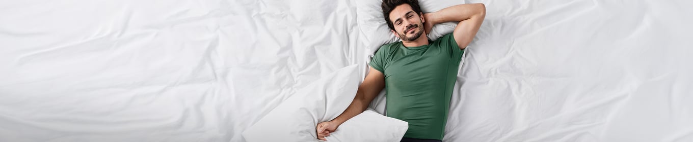 Ein Mann liegt auf einer weißen Decke und Kissen. Er hält ein weißes Kissen in den Händen und trägt ein grünes T-Shirt von CALIDA.