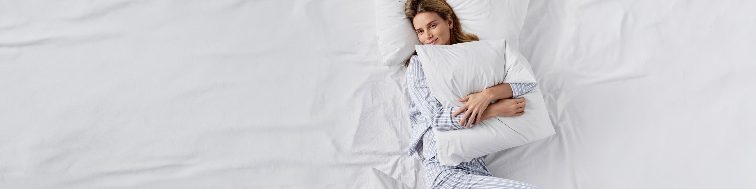 A woman is lying on a white blanket and pillow. She is holding a white pillow in her hands and wearing blue-and-white-striped CALIDA pajamas.