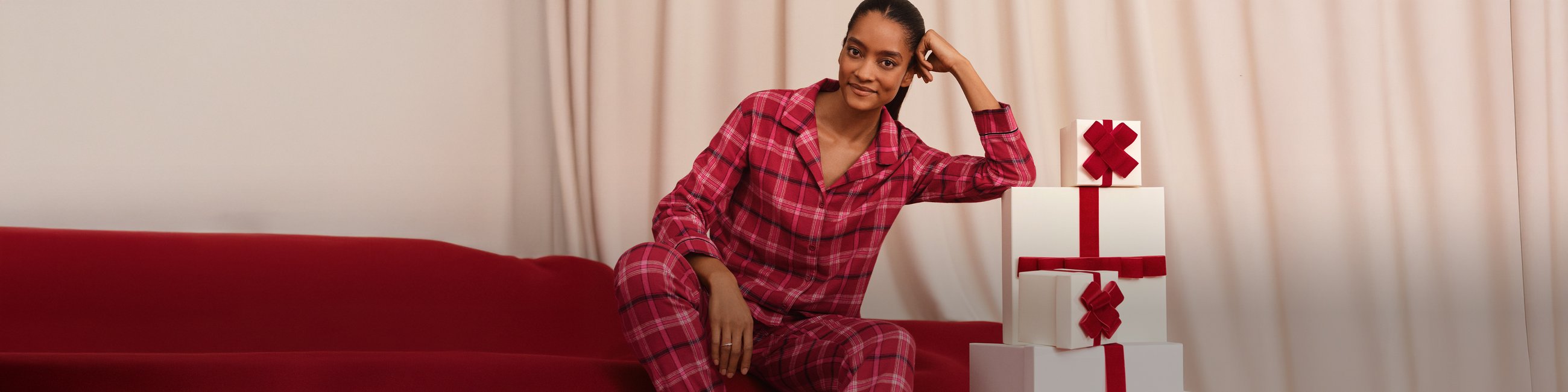 Das Bild zeigt eine Dame im roten Flanellpyjama vor einem Berg an Geschenken