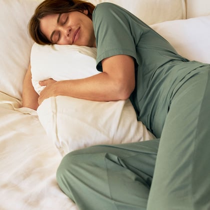 The model is lying in a bed and is wearing balancing nightwear in green.