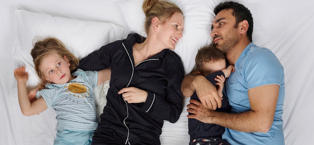 Family lying together on a white bed; two adults and two children wearing CALIDA sleepwear, relaxed bedtime scene illustrating why sleeping in pyjamas is beneficial.