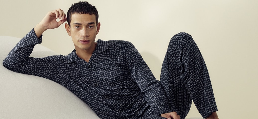 Man sitting on a soft surface. His right arm is bent so that he is touching his hair with his fingers. He is wearing Calida nightwear from the 'Relax Selected' range