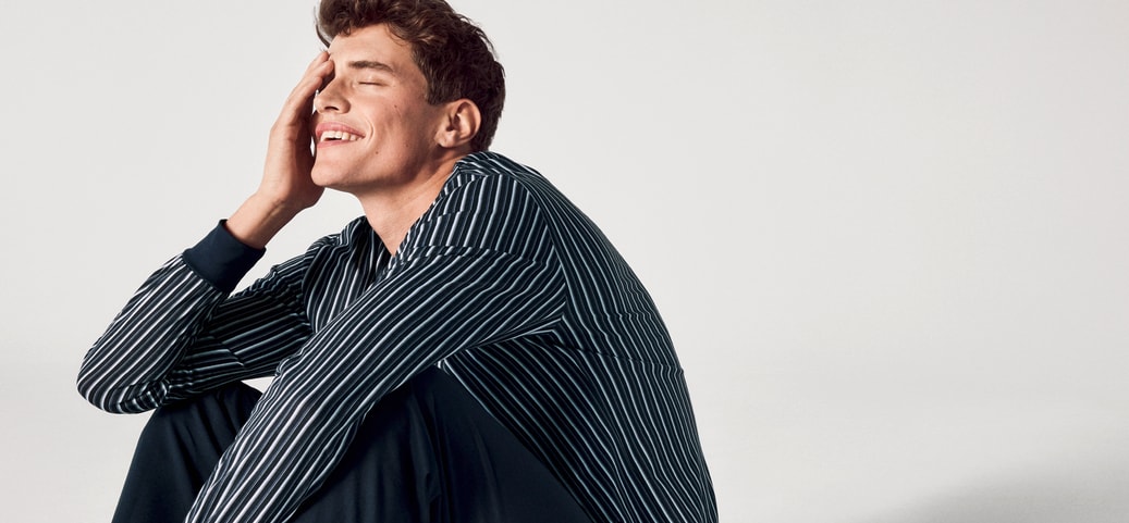 A man in striped CALIDA pyjamas sits on the floor smiling and holding a hand to his face with his eyes closed, looking relaxed and content.