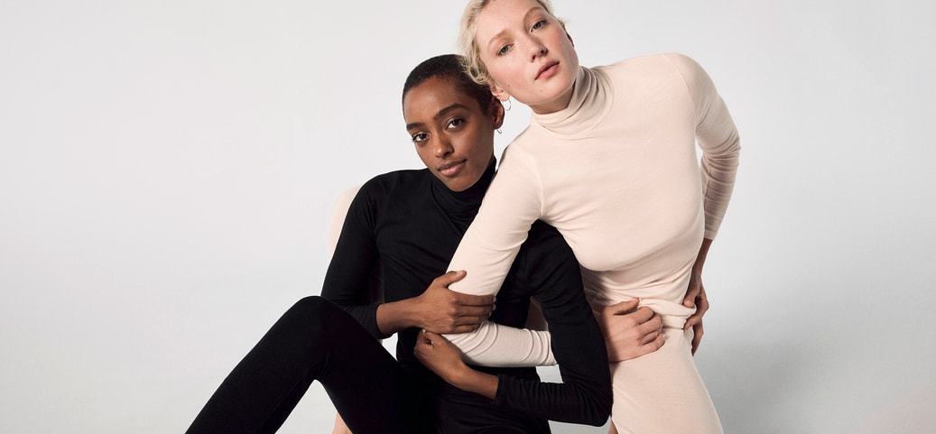 Two people leaning closely together in black and beige turtleneck outfits against a light background.