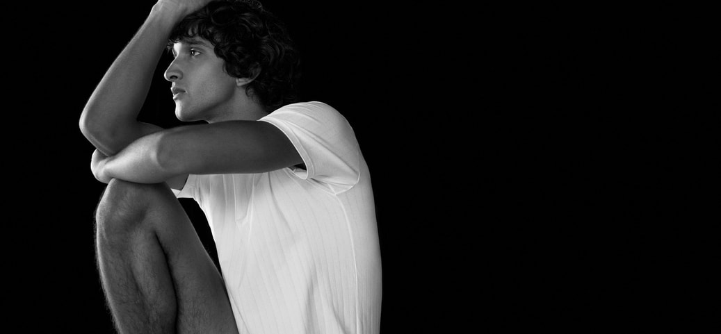 Person in a white T‑shirt sitting thoughtfully against a black background – minimalist, contemplative portrait scene