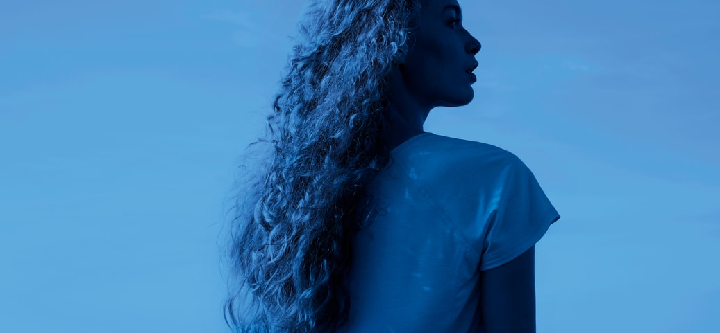 Une femme aux cheveux longs et bouclés portant un vêtement de nuit CALIDA léger, regardant la mer dans un cadre de nuit bleu profond et frais.