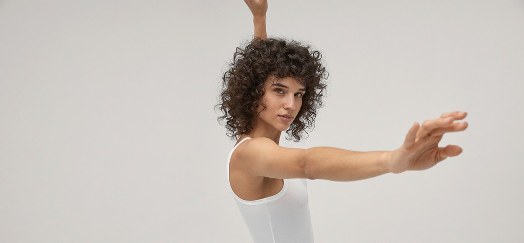 Person in einem weißen Body, seitlich stehend, mit ausgestreckten Armen vor hellem Hintergrund.