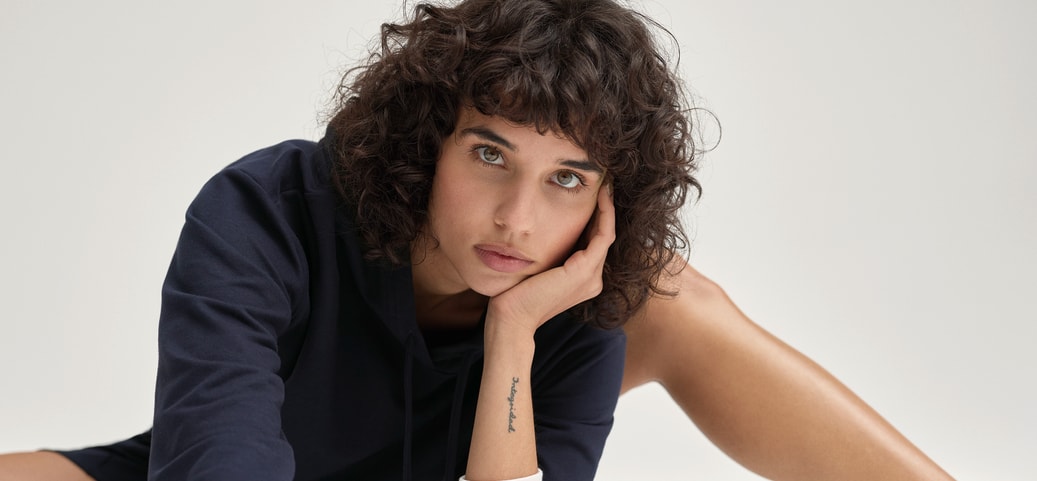Person sitting relaxed on a white surface wearing a navy long‑sleeve top with striped cuffs – minimalist, calm fashion shot