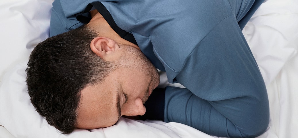 Man sleeping on his side on a white bed, wearing blue CALIDA sleepwear; symbolic image for restorative REM sleep.