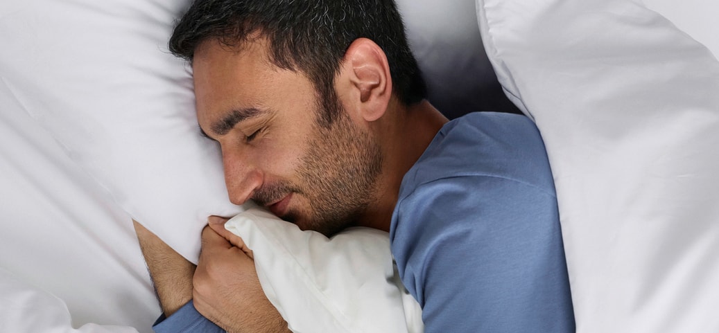 Schlafender Mann in blauer Nachtwäsche in weissem Bett; Symbolbild für erholsamen Schlaf und ein starkes Immunsystem