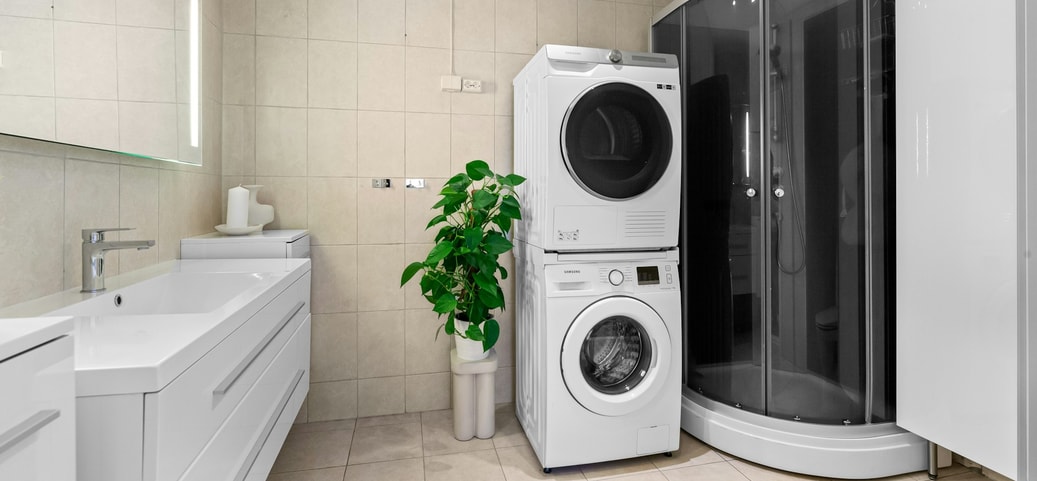 Bagno moderno con lavandino, specchio illuminato, lavatrice e asciugatrice impilate, doccia in vetro e grande vaso di piante su piastrelle chiare